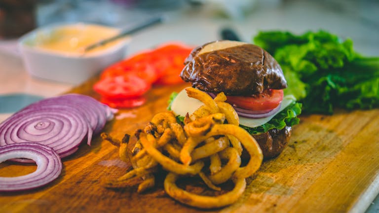 Close-up of a gourmet burger with curly fries, fresh vegetables, and ingredients on a wooden board.