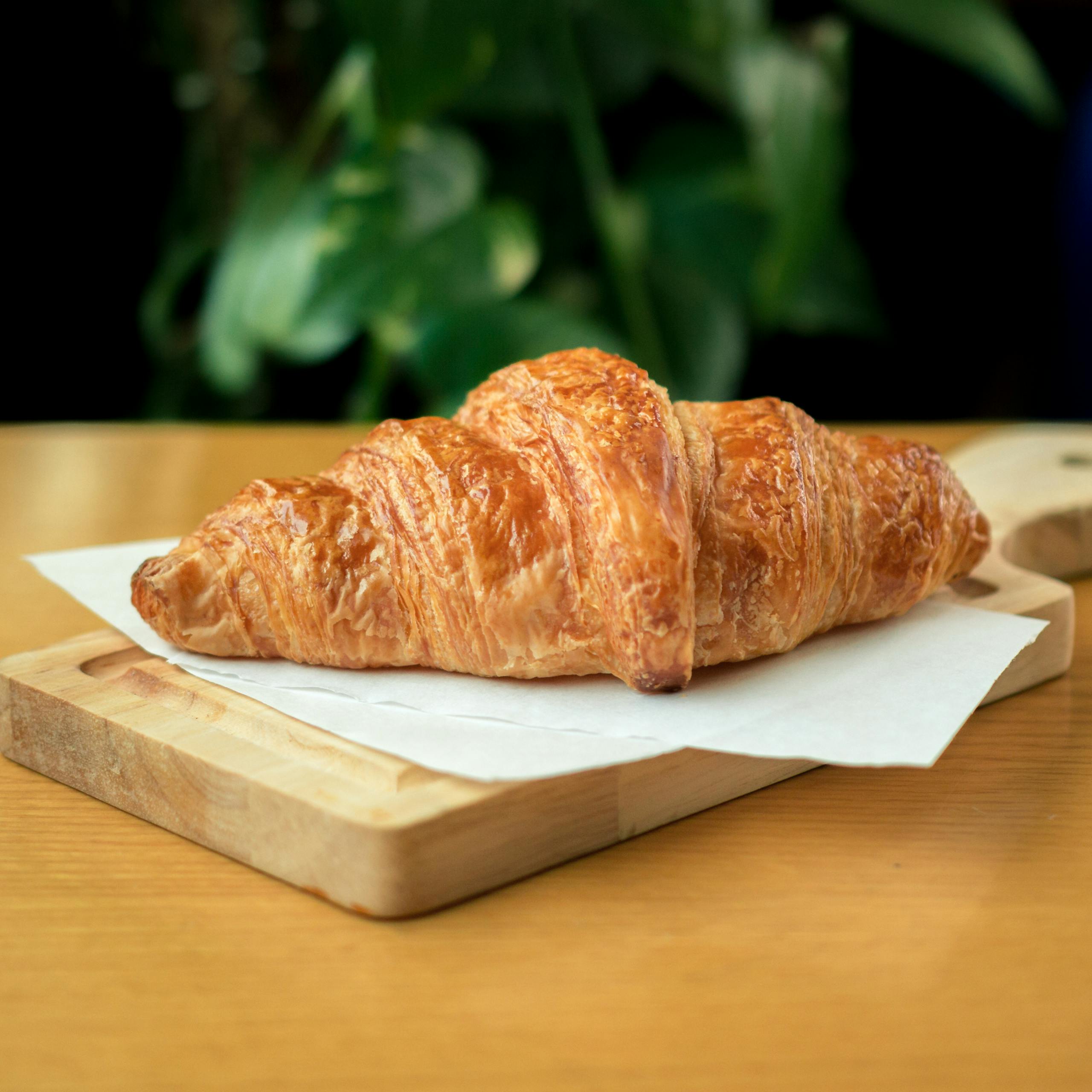 Golden-brown croissant placed on a wooden board inside a café, perfect for breakfast or snack time.